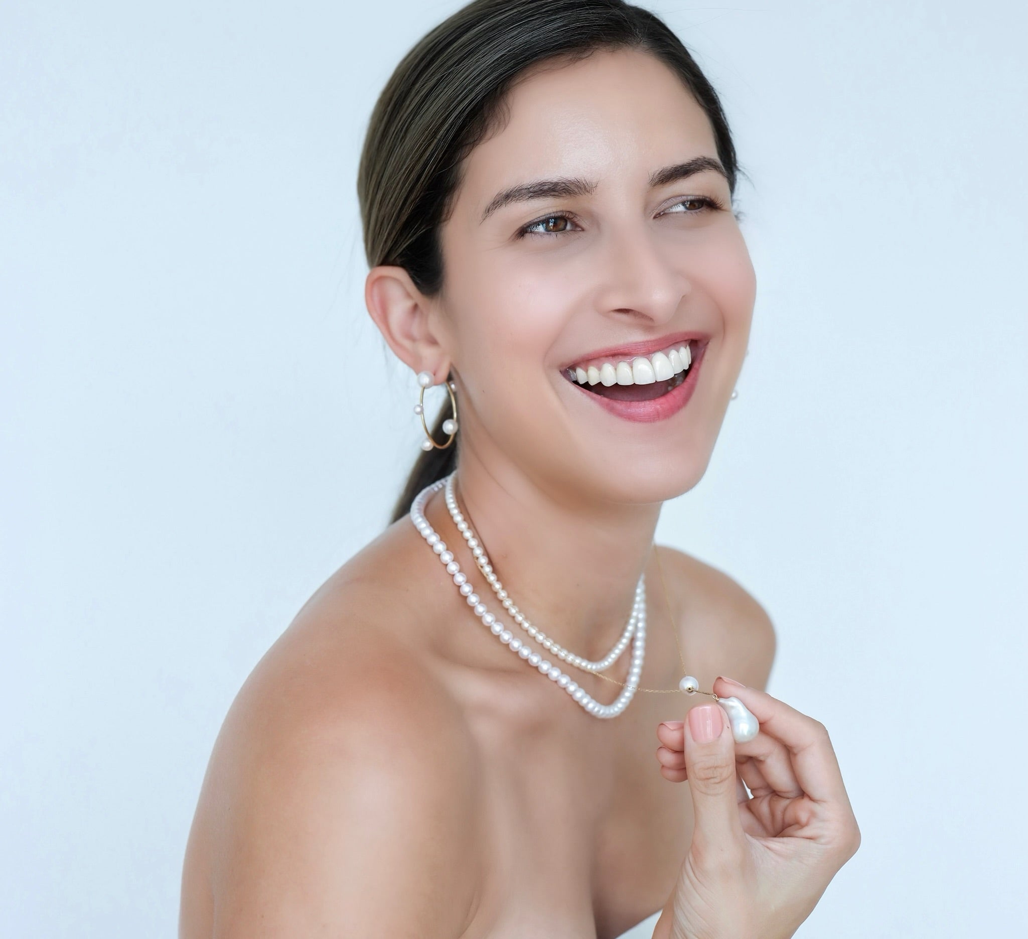 Woman wearing Akoya Hoop Earrings on ear paired with Akoya Baby Pearl Classic Necklace while holding a Baroque Pearl Charm hooked onto the necklace