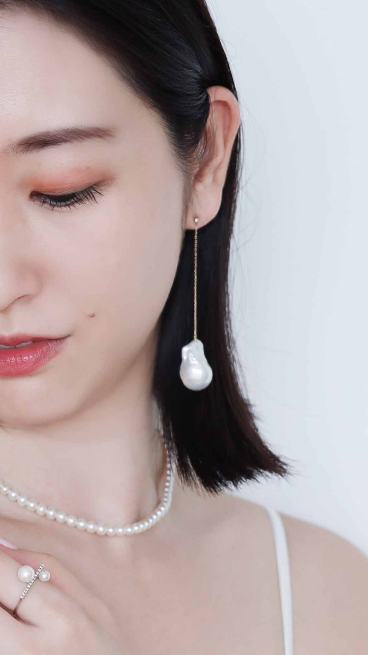 Half-faced asian woman wearing Baroque Pearl Swing Earrings paired with Akoya Baby Pearl Classic Necklace and Akoya Diamond Bar ring on left index finger